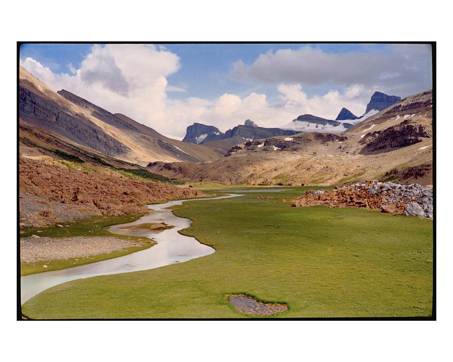 A winding river flows through a green valley surrounded by mountains with patches of snow under a cloudy sky.