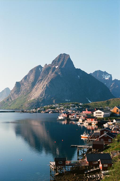 A small fishing village with red and white houses is nestled at the base of a large mountain, with its reflection visible in the calm water.