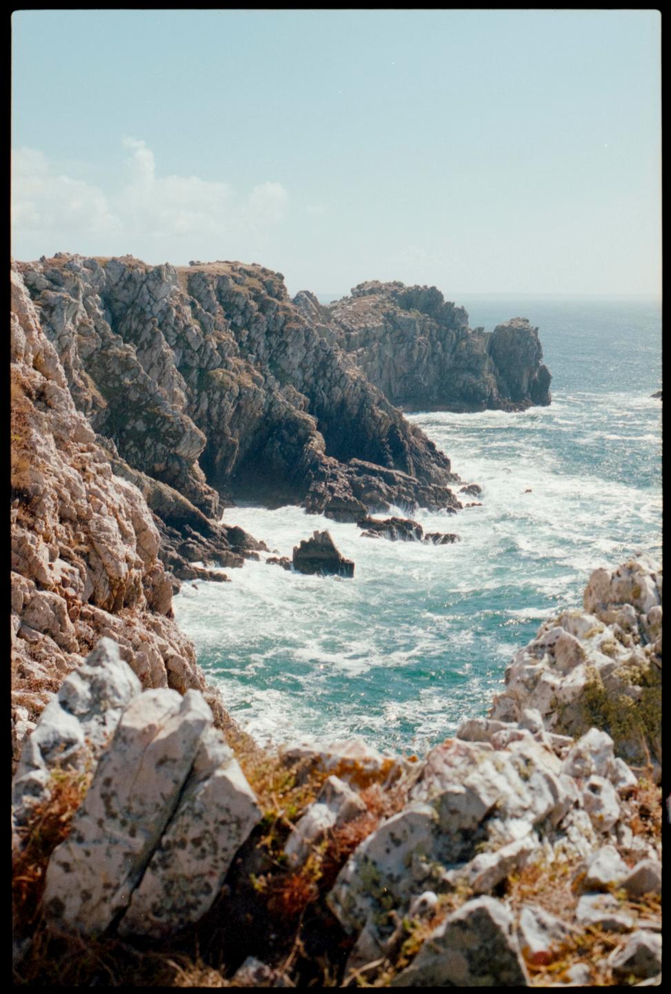 A rocky coastline with waves crashing against the shore under a clear sky.