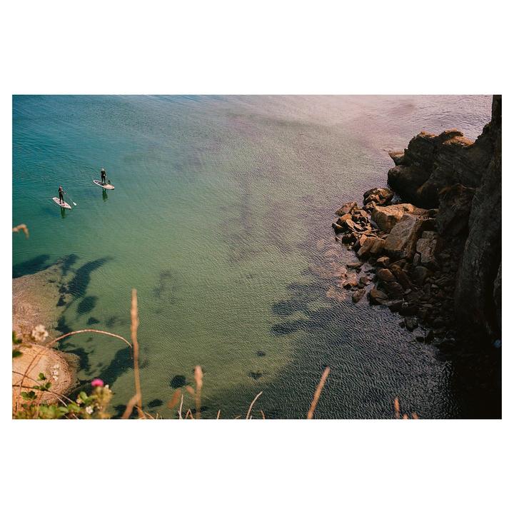 Two people paddleboard on clear water next to a rocky cliff.