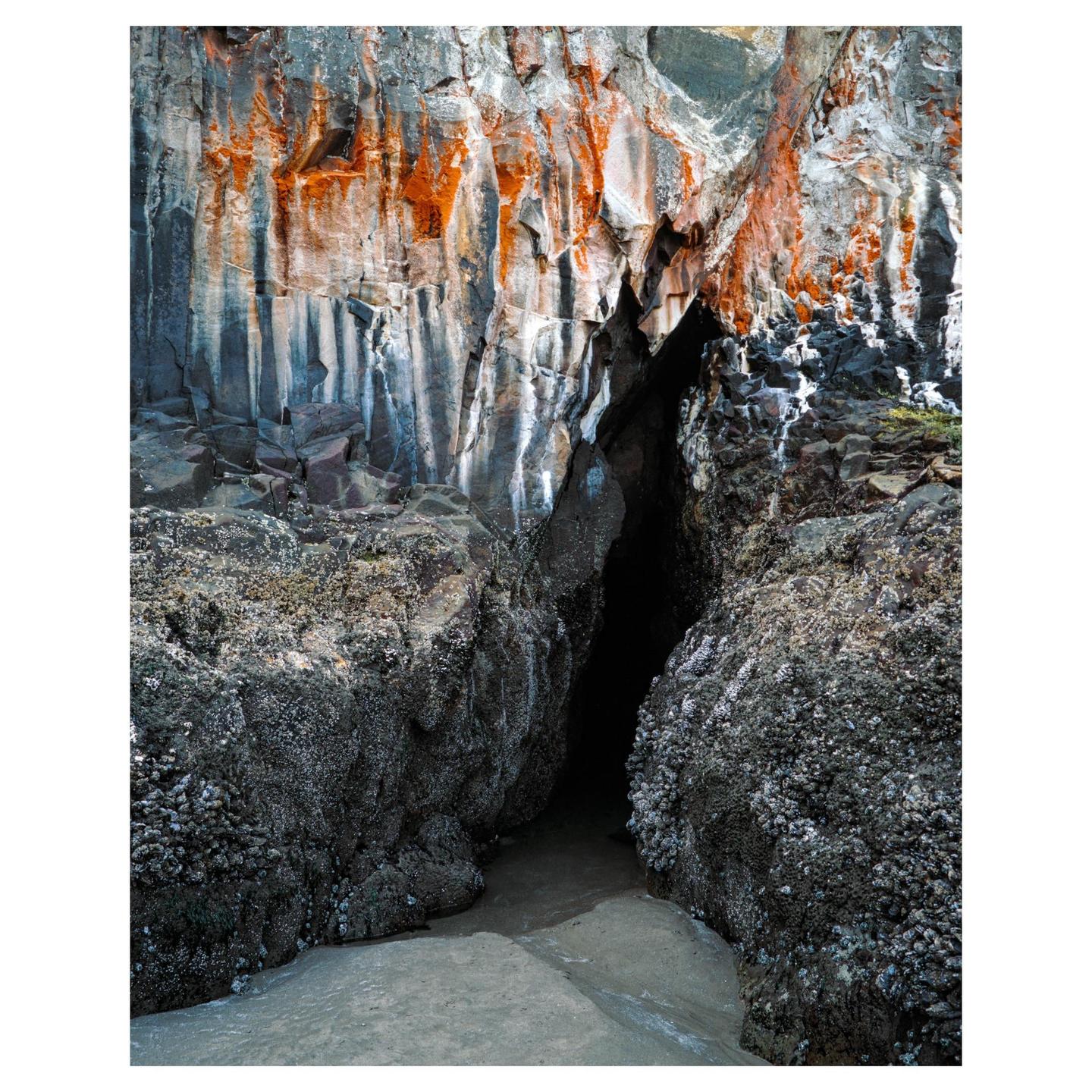 A dark cave entrance is visible between two large rocks covered in barnacles, with a cliff face above showing orange and white mineral streaks.