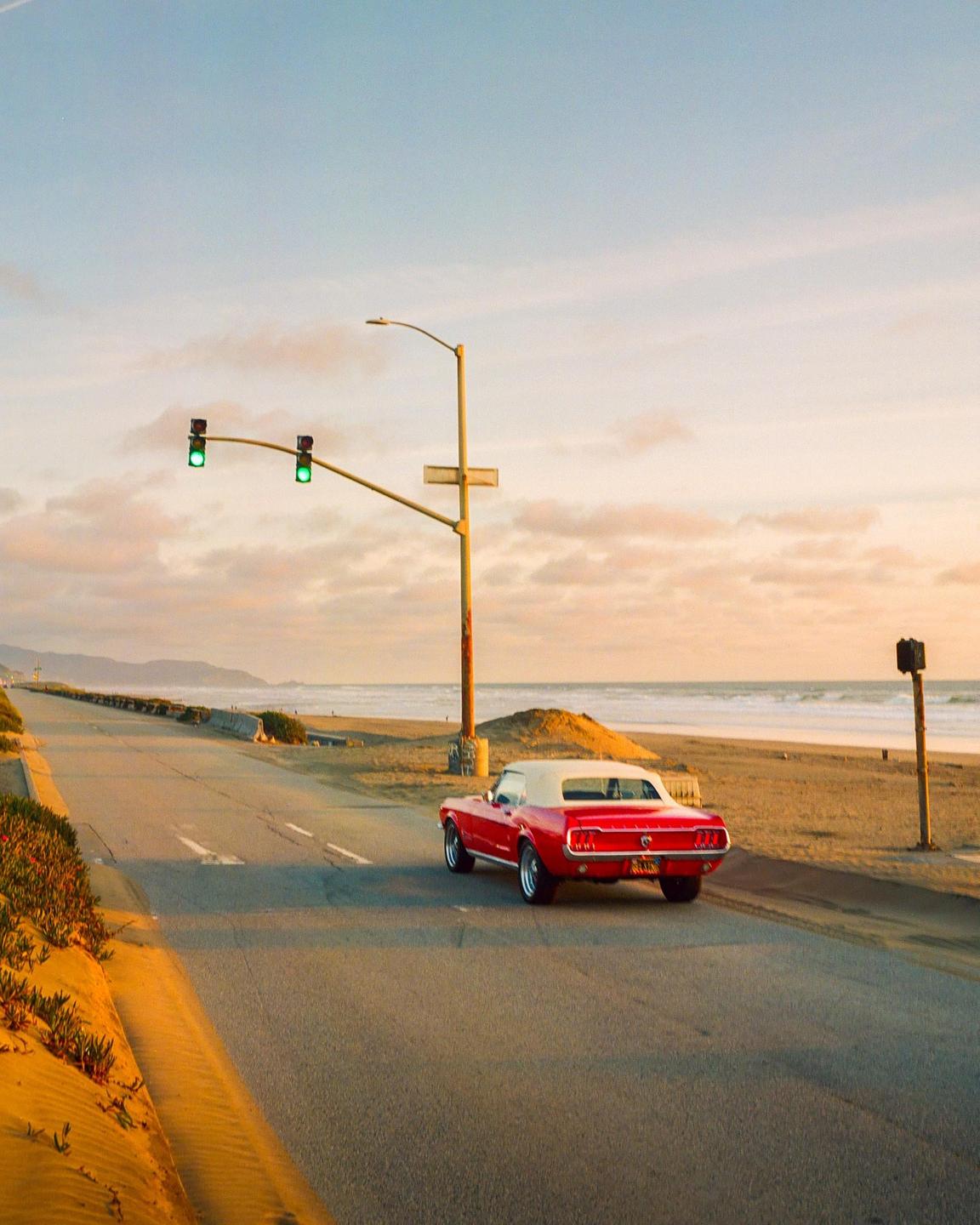 A red convertible car is driving on a road next to a beach at sunset.