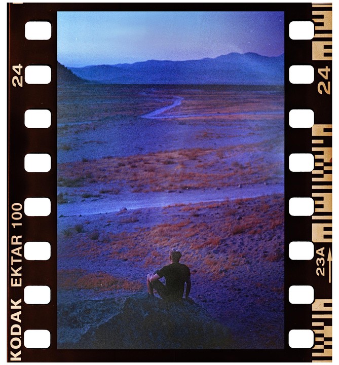 A man sits on a rock overlooking a vast, purple-hued landscape with mountains in the distance.