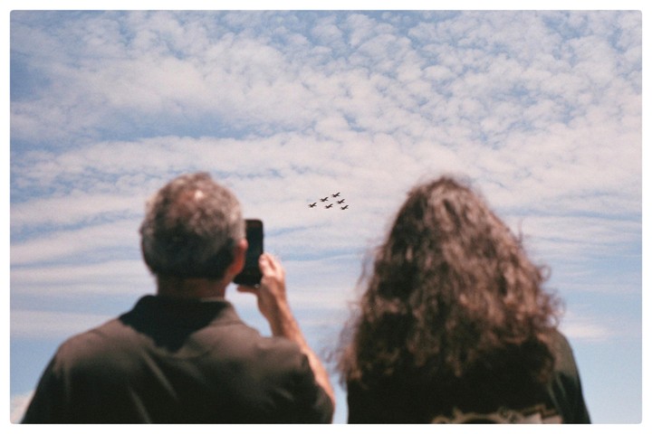 A man and a woman watch six jets flying in formation across a cloudy sky, with the man holding a phone up to record them.