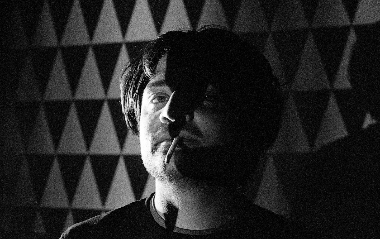 A man with a cigarette in his mouth is partially illuminated by light against a patterned wall.