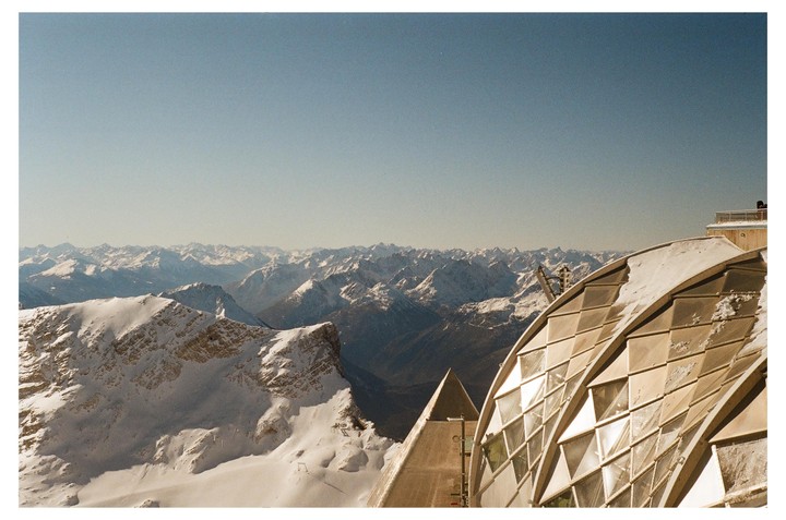 A modern building with a curved, gridded roof stands in the foreground, overlooking a vast expanse of snow-covered mountains under a clear sky.