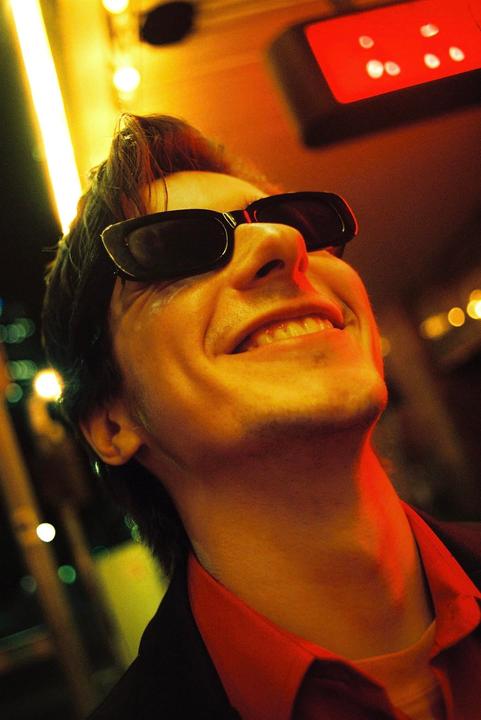 A man wearing sunglasses and a red shirt smiles while looking up, illuminated by warm light.