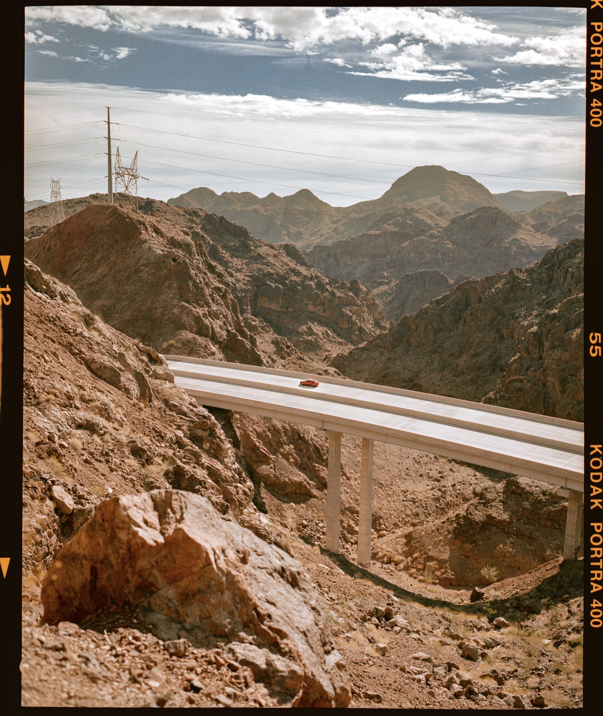 A red car drives on a highway bridge over a desert canyon with mountains in the background.