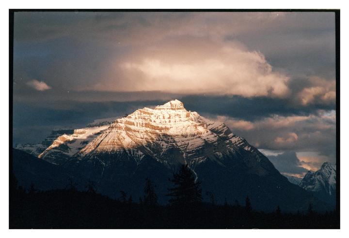 A snow-capped mountain peak is illuminated by a ray of sunlight under a cloudy sky.