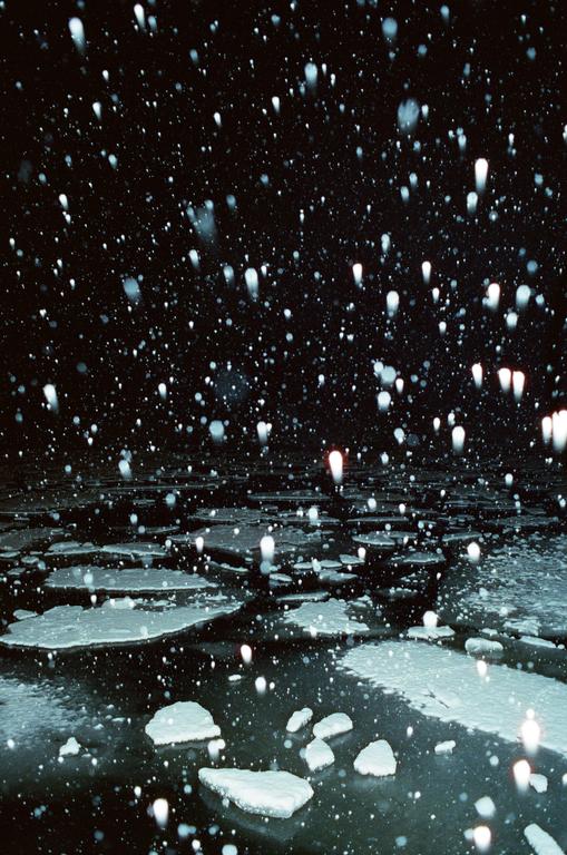 Snow falls over a body of water with floating ice at night.