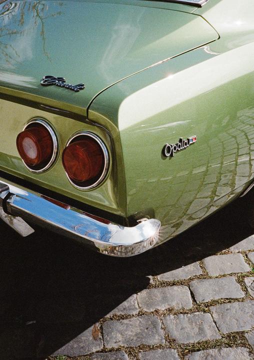 The rear of a green vintage car with round taillights and a chrome bumper is parked on a cobblestone street.