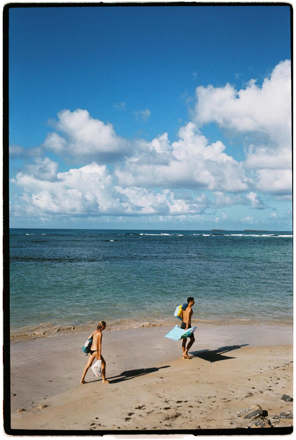 A couple walks along a sandy beach next to the ocean under a blue sky with clouds.
