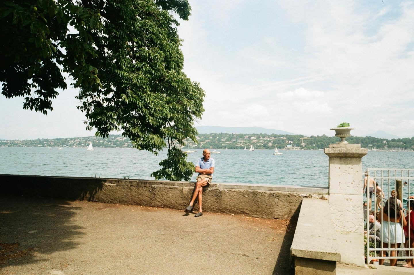A man sits on a stone wall by a lake with sailboats and distant hills under a partly cloudy sky.