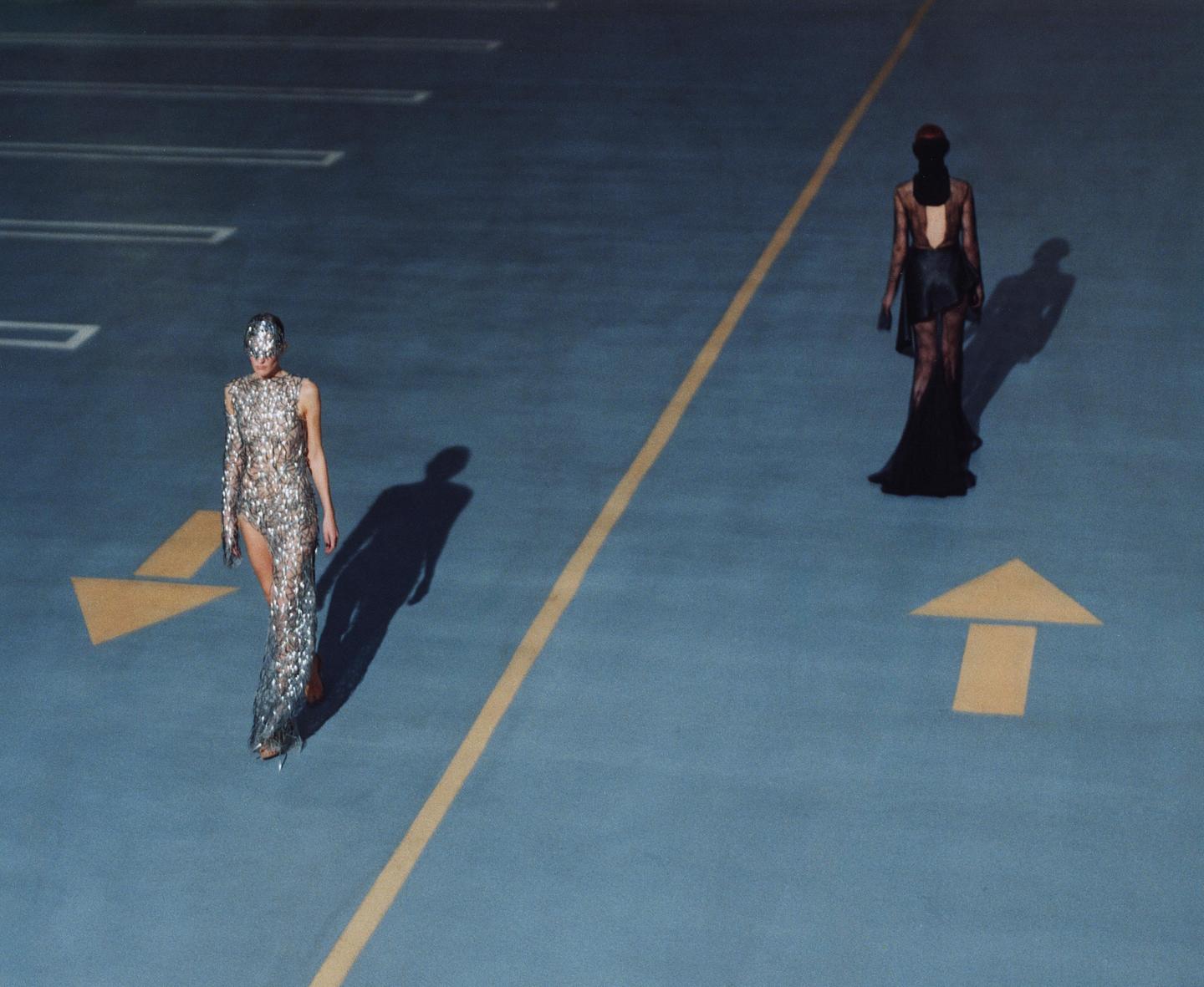 Two women in elaborate dresses walk on a blue asphalt surface with yellow lines and arrows.