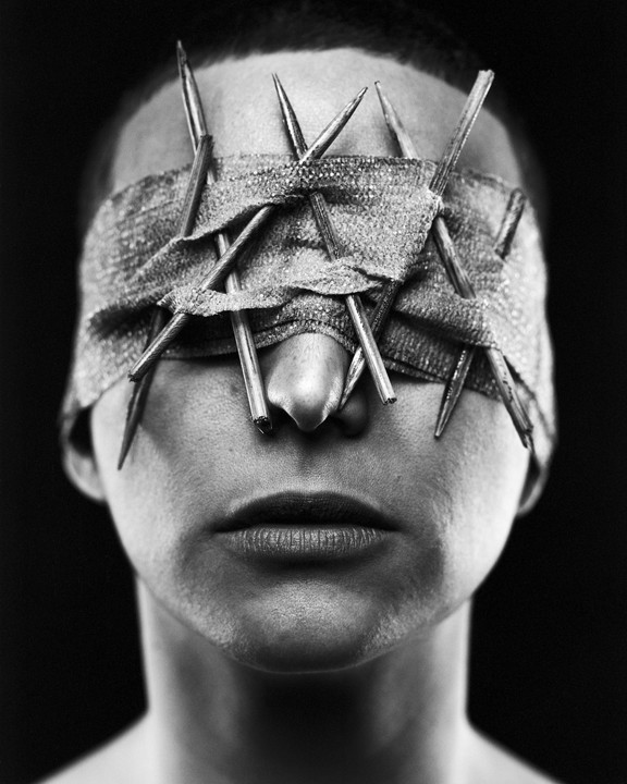 A person with a blindfold made of fabric and sharp metal objects covering their eyes is shown in a close-up portrait.