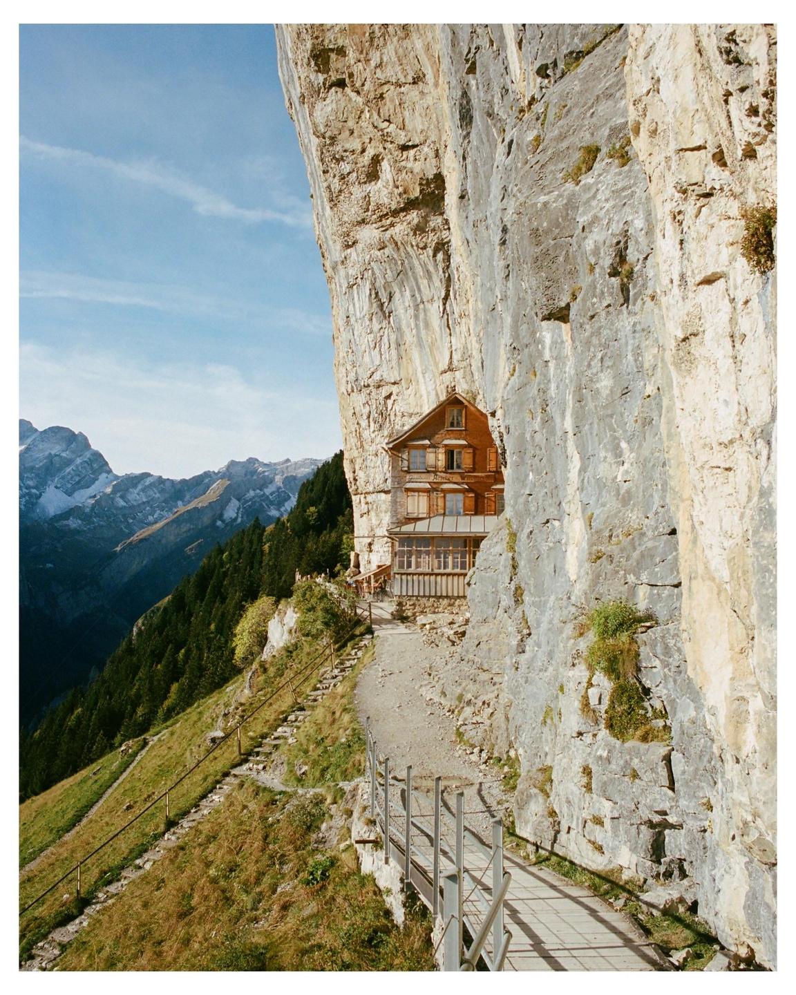 A wooden building is built into the side of a large rock cliff with a path leading to it and mountains in the background.