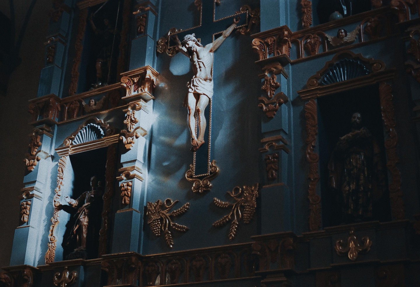 A statue of Jesus on the cross is mounted on a dark blue wall with gold accents and other statues in niches.