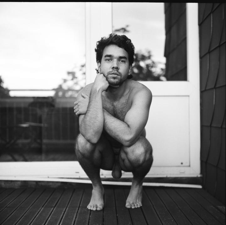 A man crouches naked on a wooden deck, looking directly at the viewer.