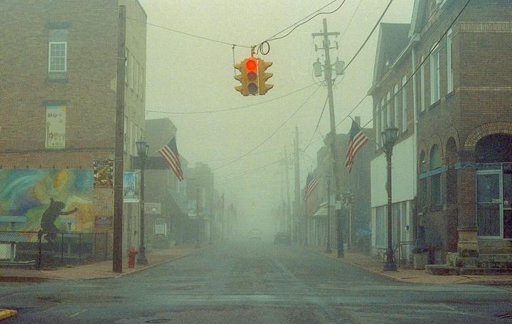 A street with buildings and American flags is shrouded in thick fog, with a red traffic light hanging overhead.