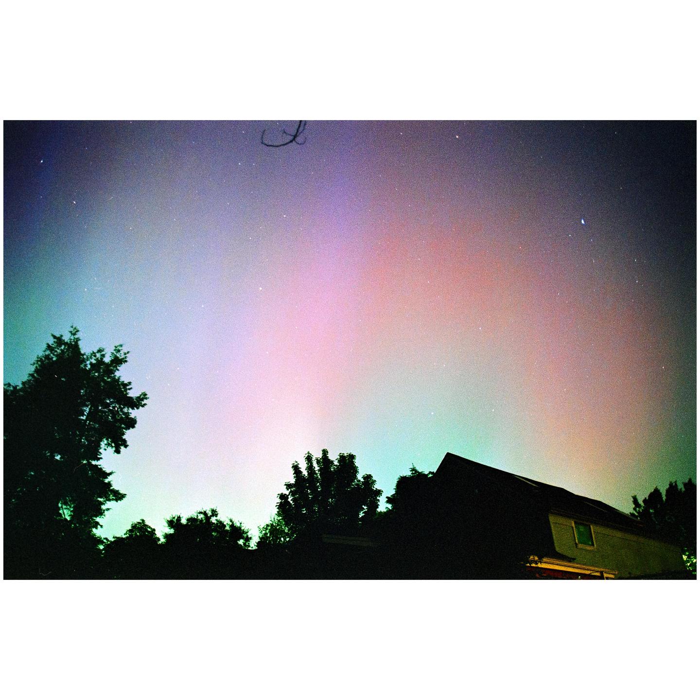The aurora borealis glows in the night sky above a house and trees.
