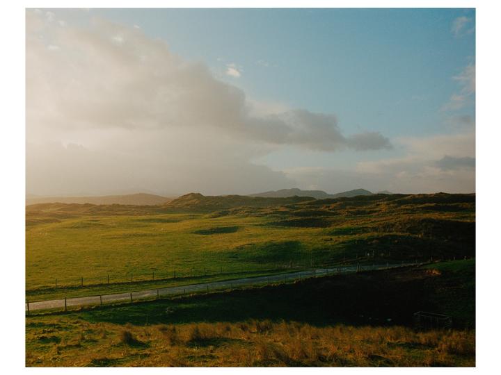 A road runs through a green, hilly landscape under a partly cloudy sky.