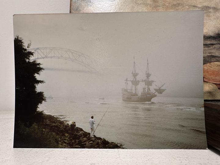 A man fishes from a rocky shore as a tall ship sails past a bridge in the fog.