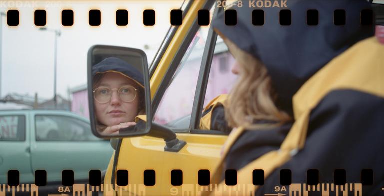 A woman in a yellow jacket and blue hat is reflected in the side mirror of a yellow vehicle, with another car visible in the background.