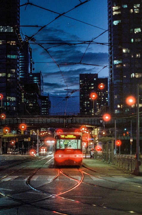 A red streetcar is stopped on tracks in a city at night, surrounded by tall buildings and overhead wires.