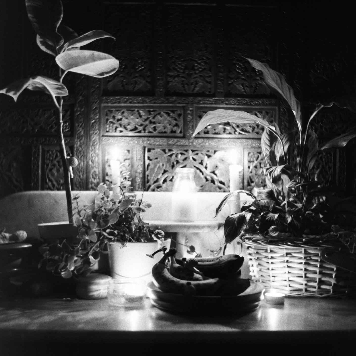 A still life shows potted plants, bananas on a plate, and lit candles in front of an ornate wooden screen.