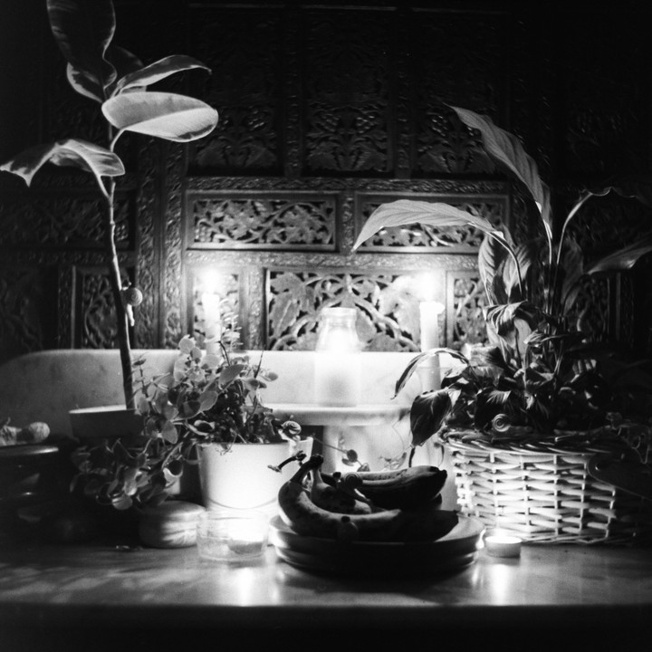 A still life shows potted plants, bananas on a plate, and lit candles in front of an ornate wooden screen.
