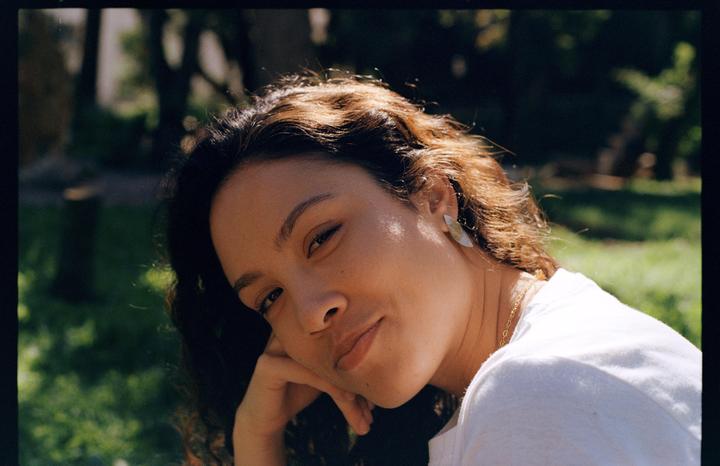 A woman with curly hair and a white shirt is resting her head on her hand and looking at the viewer.