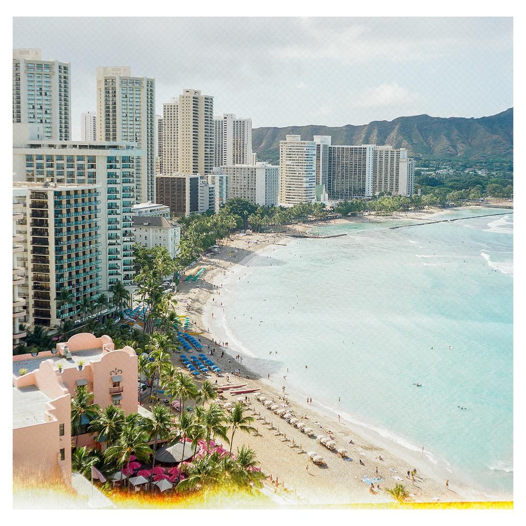 A high-angle view shows a long beach with many people, palm trees, and tall buildings next to a calm ocean.