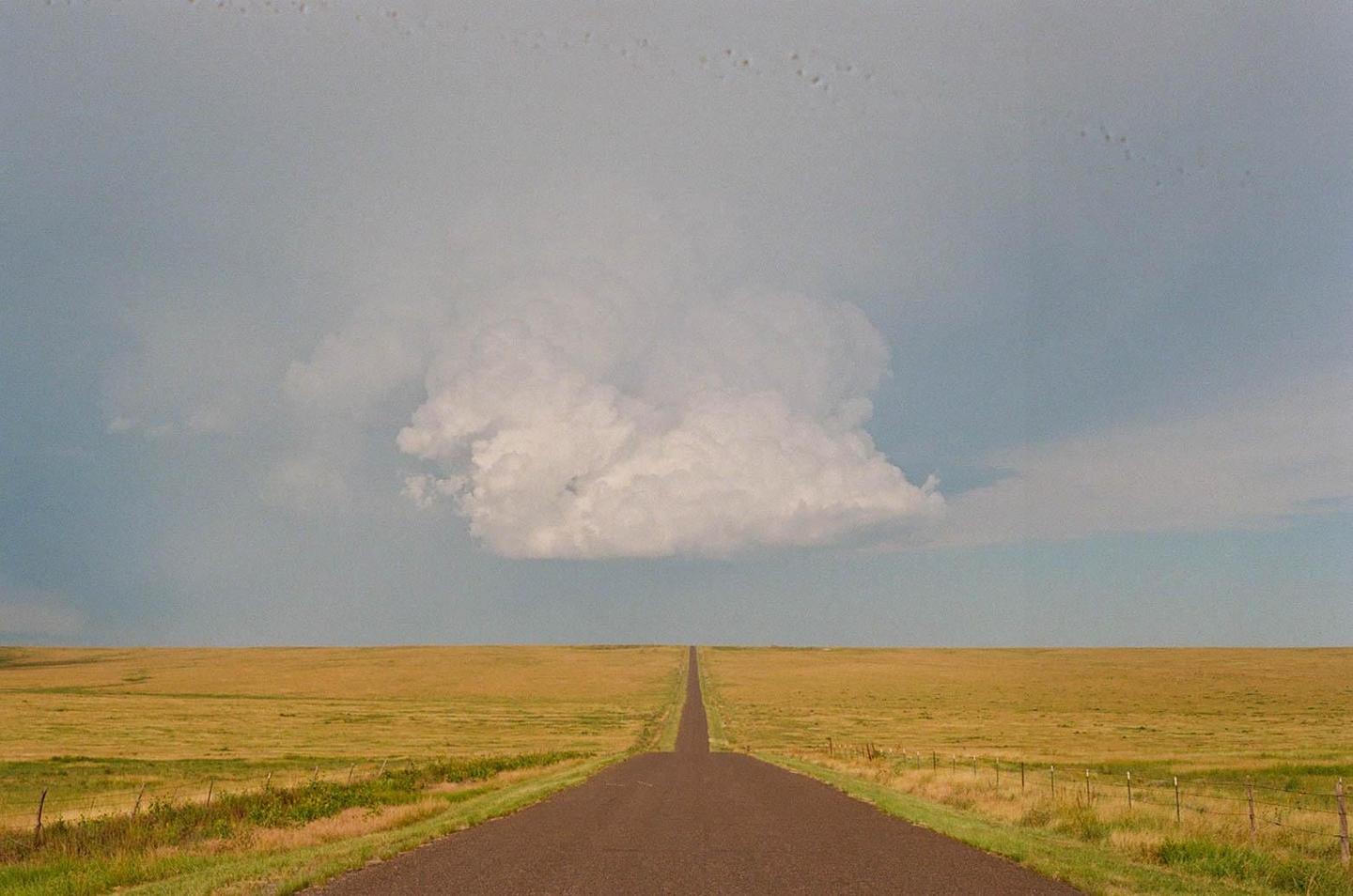 A long, straight road extends into the distance through a flat, grassy plain under a sky with a large cloud formation.