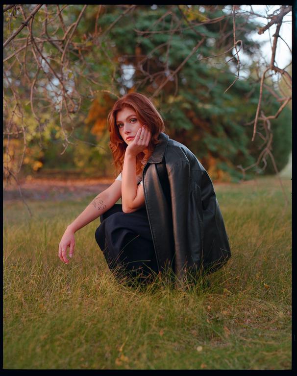 A woman with red hair crouches in a field of grass with a leather jacket draped over her shoulder.