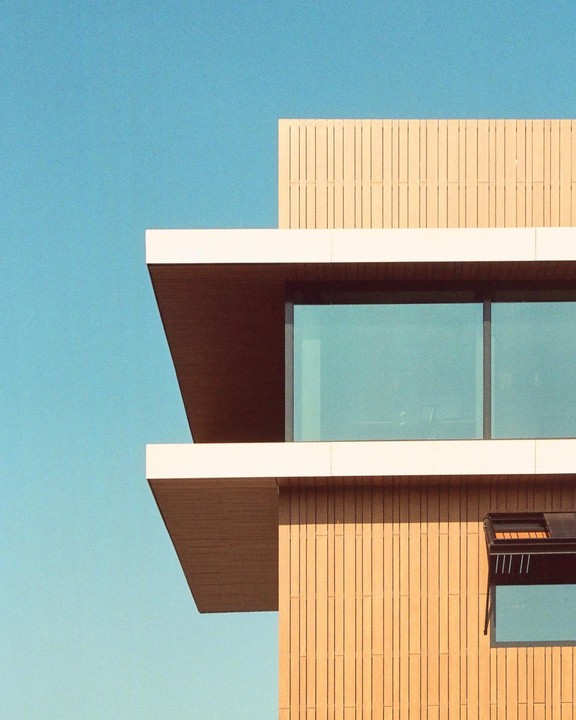 A modern building with wood paneling and large windows is set against a clear blue sky.