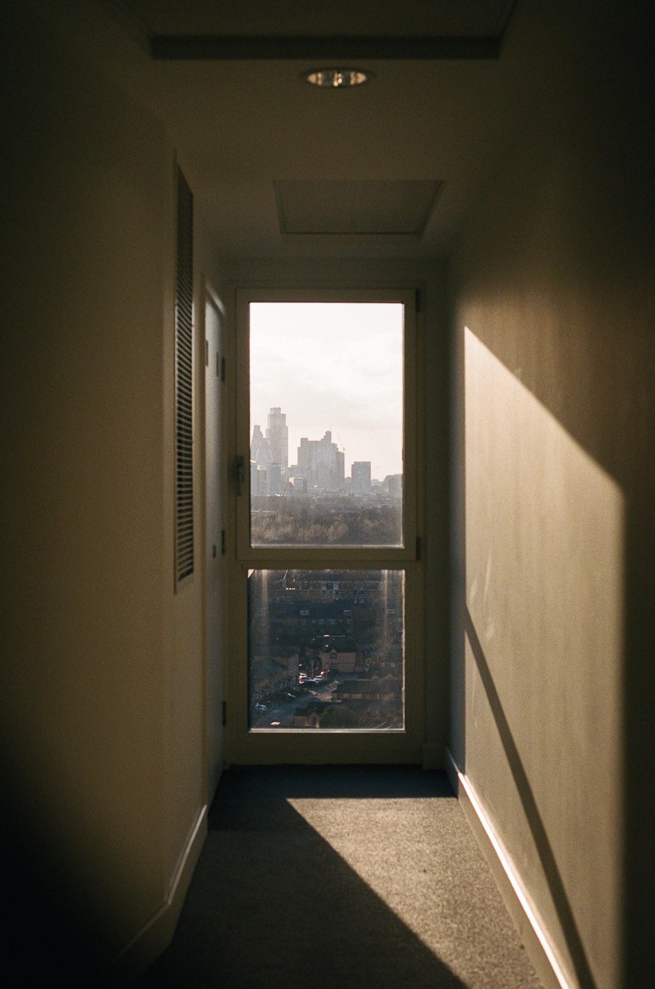 A window at the end of a hallway looks out onto a city skyline and a residential neighborhood.