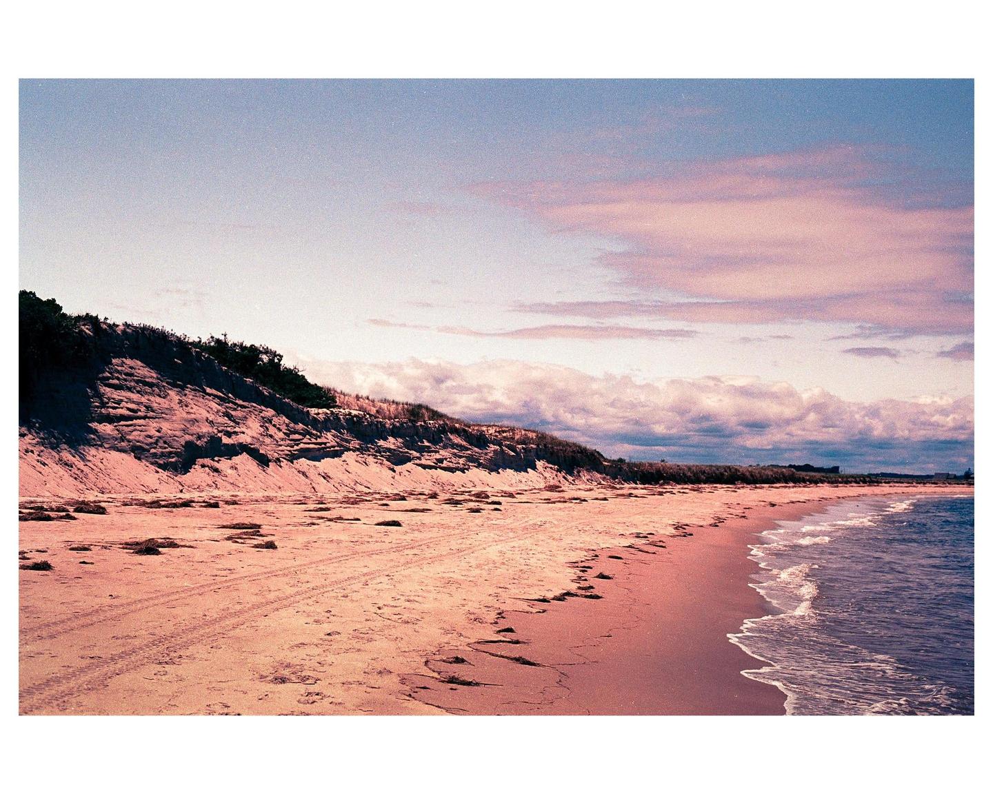 A sandy beach with a steep dune on the left and gentle waves on the right is visible under a cloudy sky.