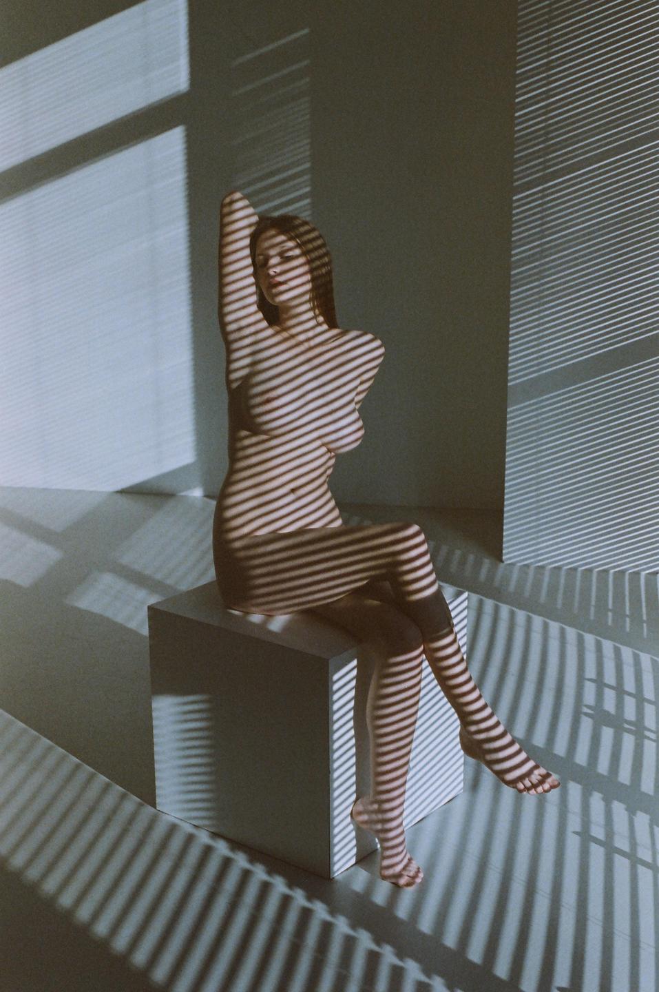 A nude woman sits on a white cube with shadow stripes cast across her body and the surrounding walls and floor.