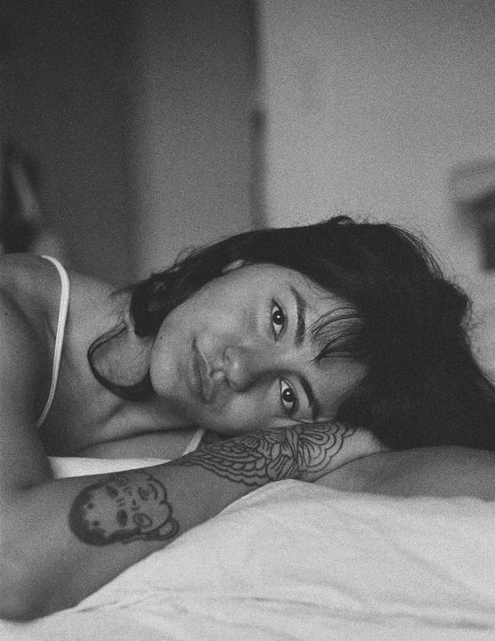 A woman with dark hair and tattoos on her arm is lying on a bed.