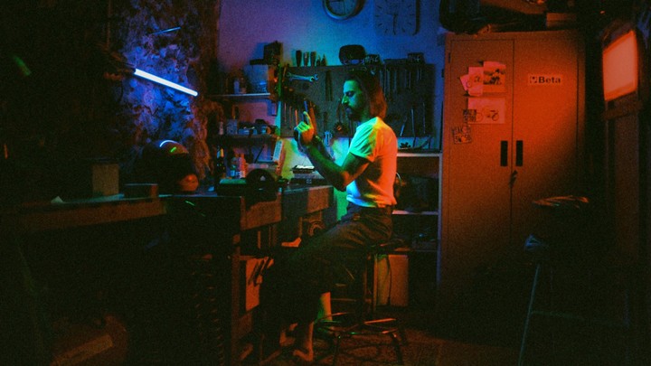 A man sits on a stool in a workshop, holding a prop weapon, illuminated by blue and red light.