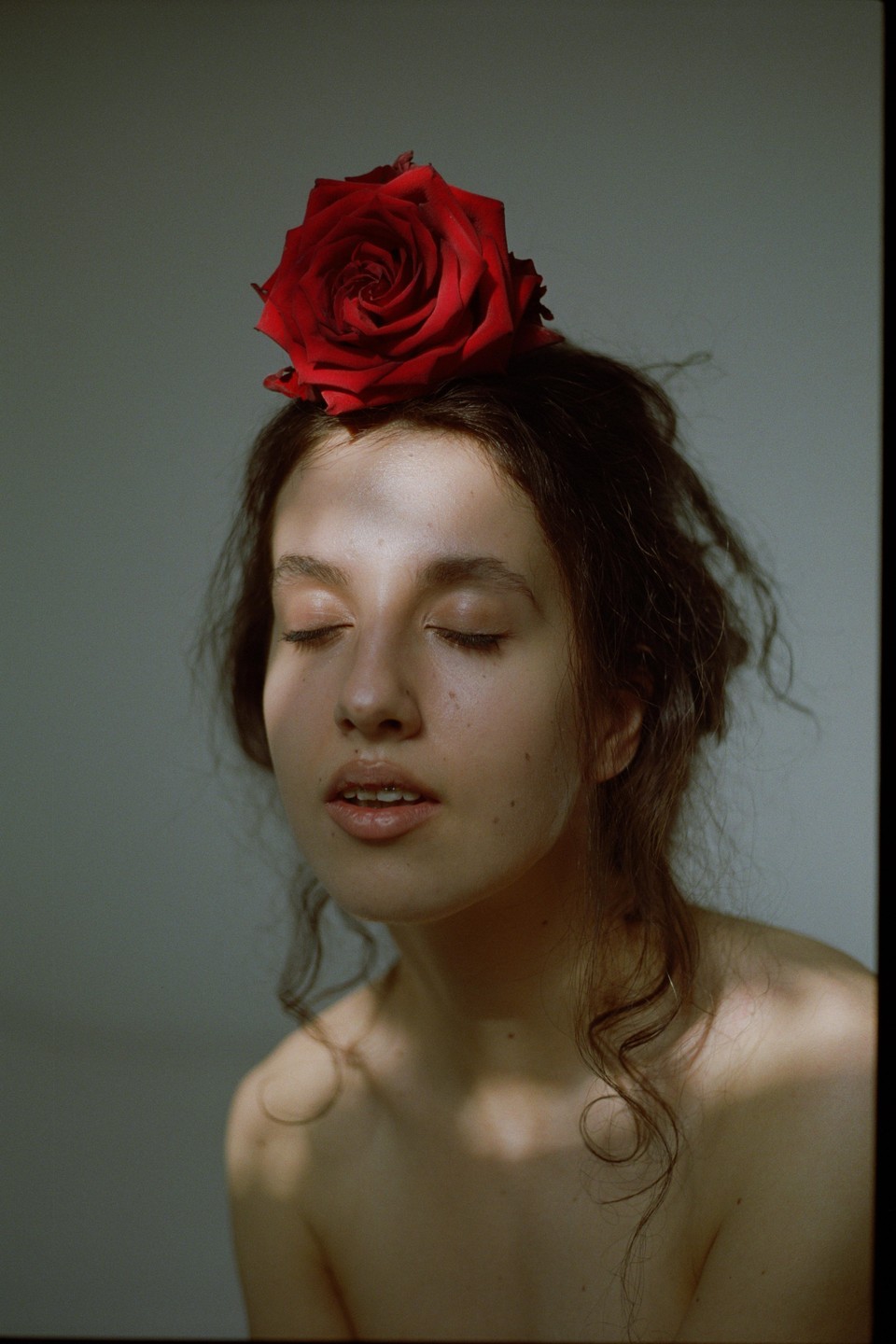 A woman with closed eyes and a red rose on her head is illuminated by a shaft of light.