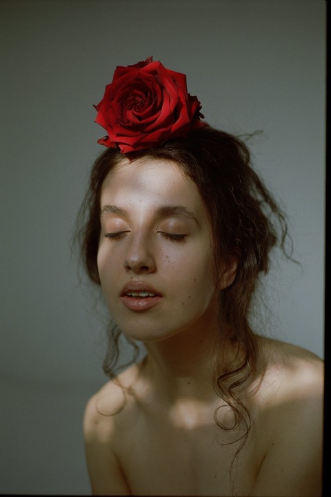A woman with closed eyes and a red rose on her head is illuminated by a shaft of light.