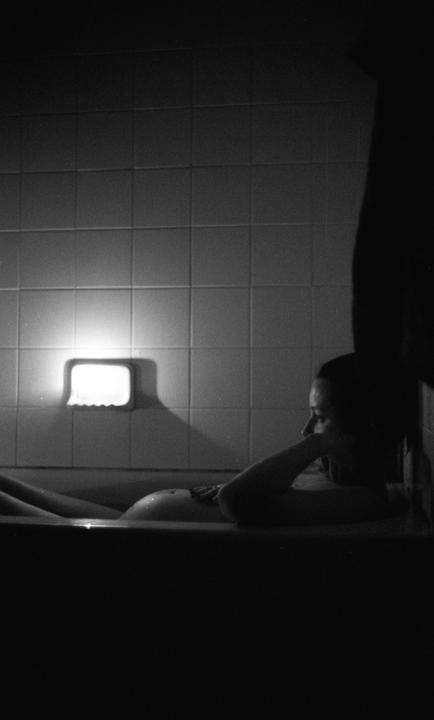 A pregnant woman is relaxing in a bathtub, illuminated by a light on the tiled wall.