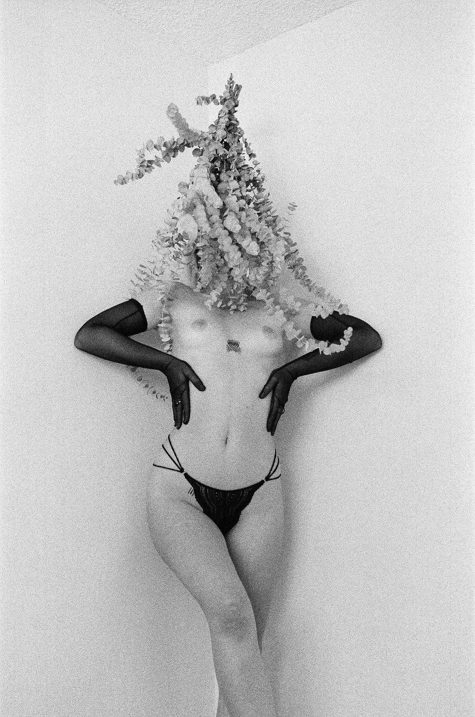 A woman with a floral arrangement covering her head and shoulders poses against a white wall.
