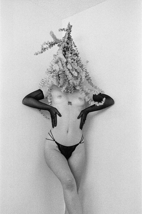 A woman with a floral arrangement covering her head and shoulders poses against a white wall.