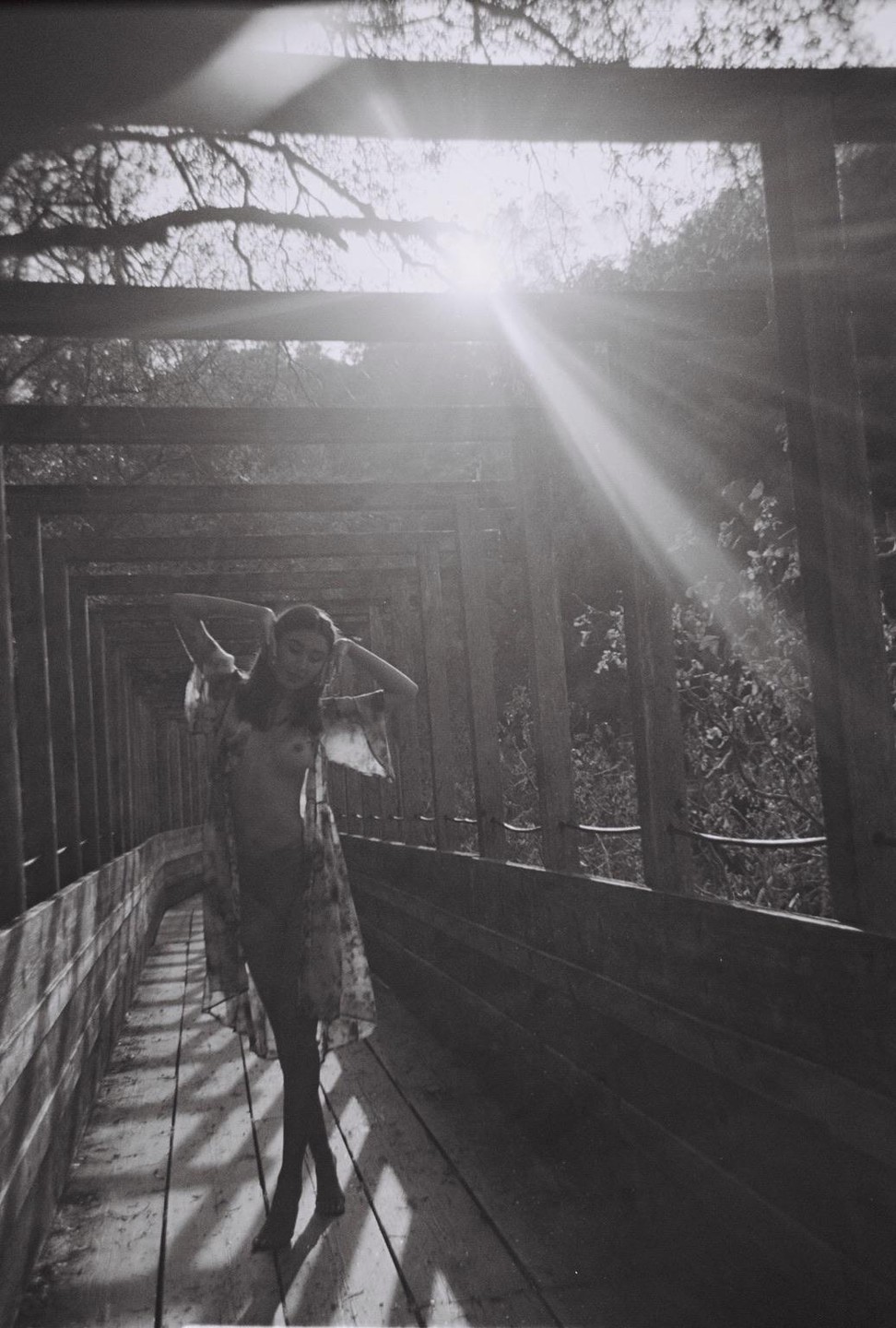 A woman stands on a wooden walkway with her arms raised, backlit by the sun.