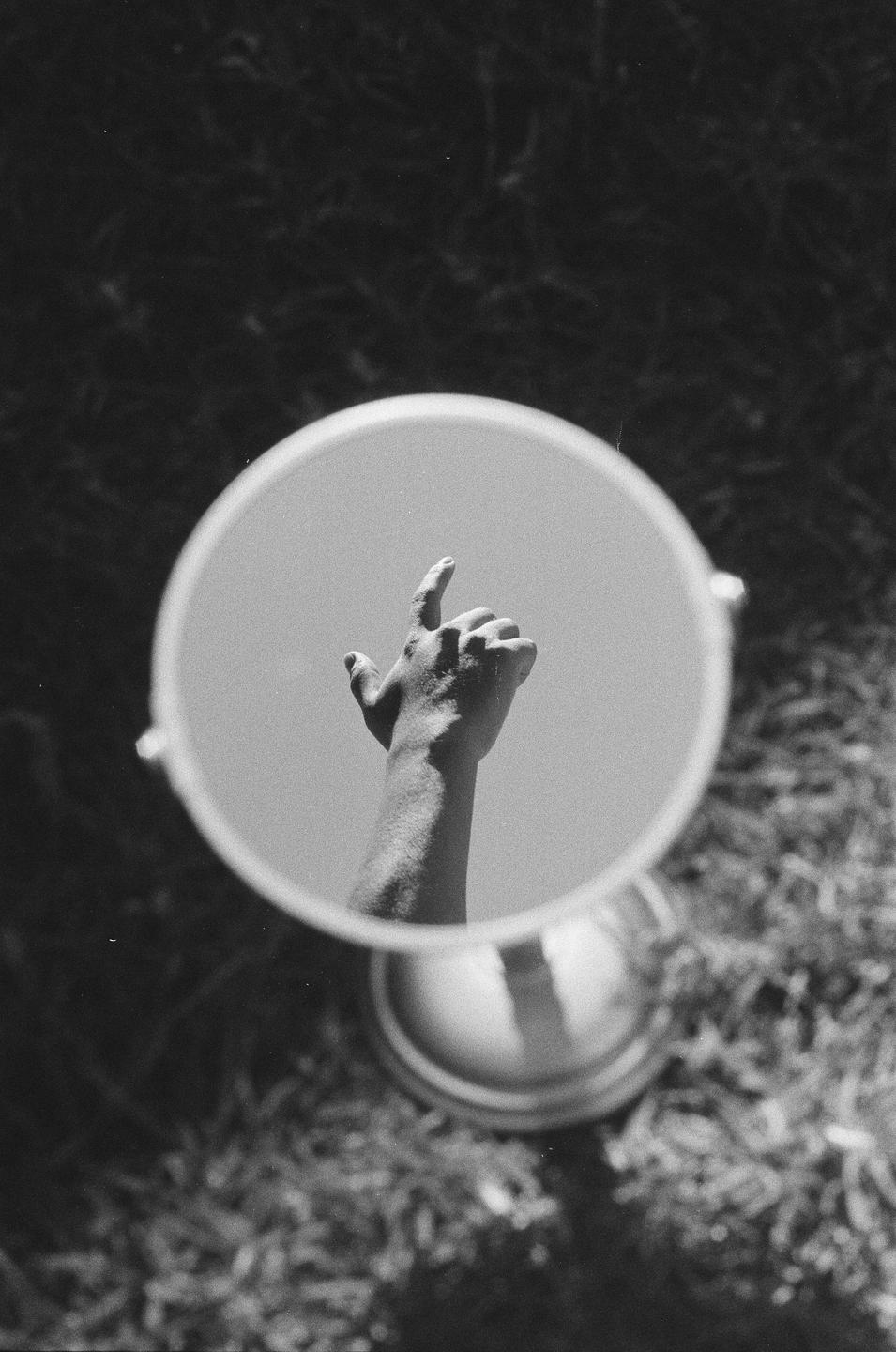 A hand is reflected in a round mirror, pointing upwards towards a bright sky.