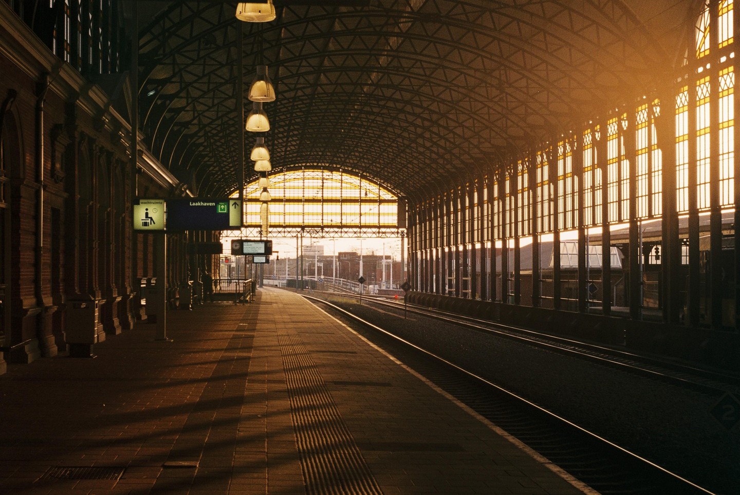 A train station platform is empty with tracks leading into the distance and warm light coming through the windows.