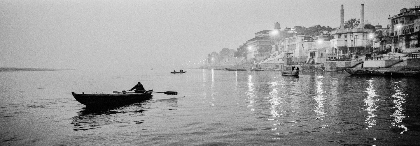 A person rows a small boat on a wide river with a city on the bank in the distance.