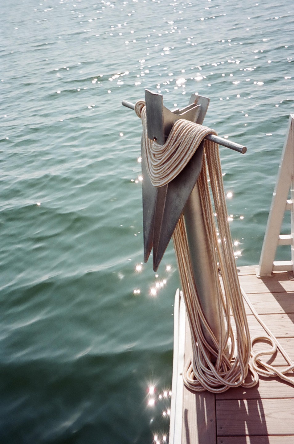 An anchor with rope wrapped around it sits on a wooden dock next to sparkling water.
